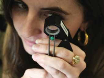 A woman uses a loupe to inspect a green gemstone.